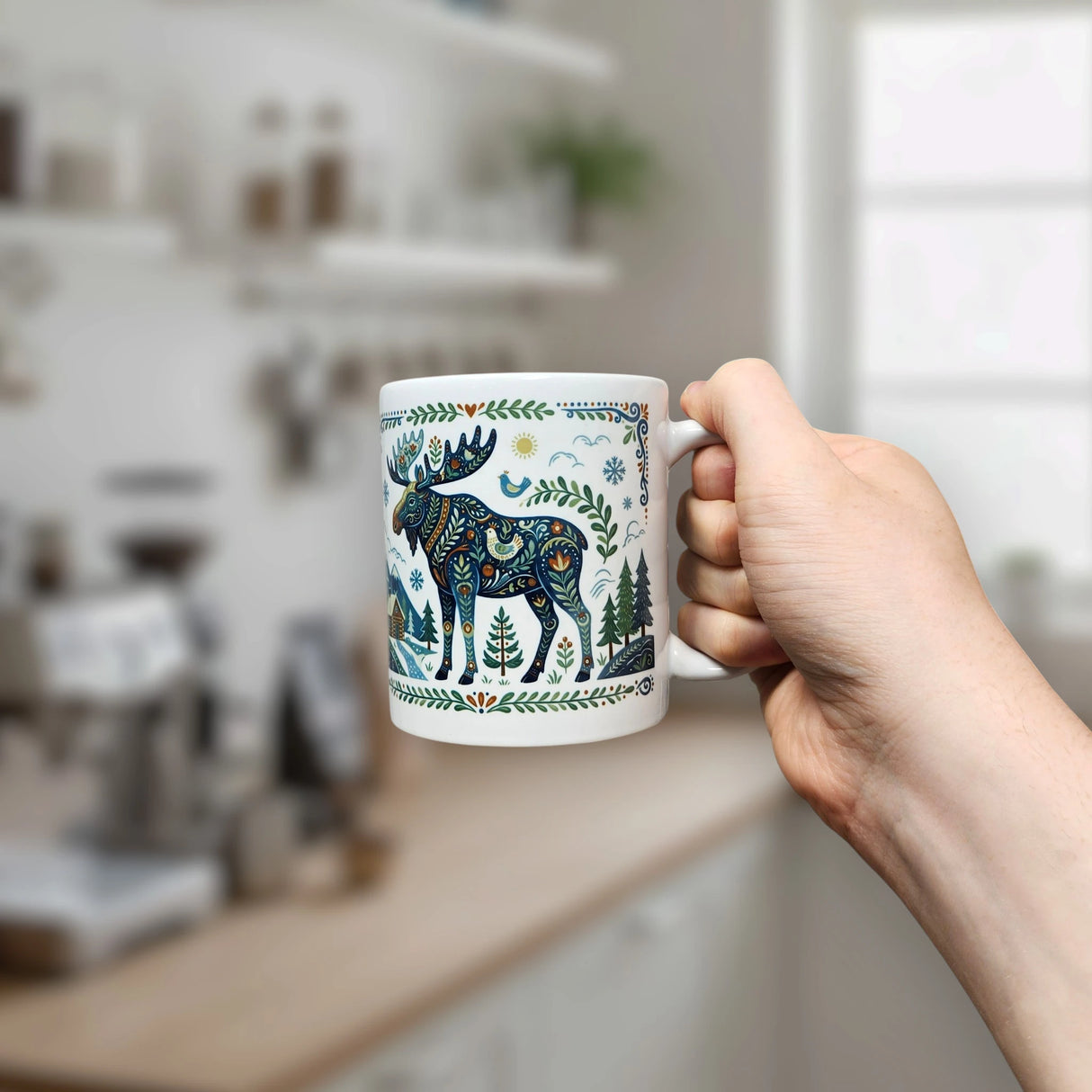 A hand holds the Nordic Wholesale "Nordic Forest Moose Ceramic Mug" (11 oz), its white ceramic surface adorned with a blue and green patterned moose and trees, with a blurred kitchen in the background.