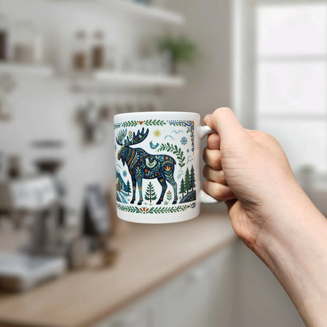 A hand holds the Nordic Wholesale "Nordic Forest Moose Ceramic Mug" (11 oz), its white ceramic surface adorned with a blue and green patterned moose and trees, with a blurred kitchen in the background.