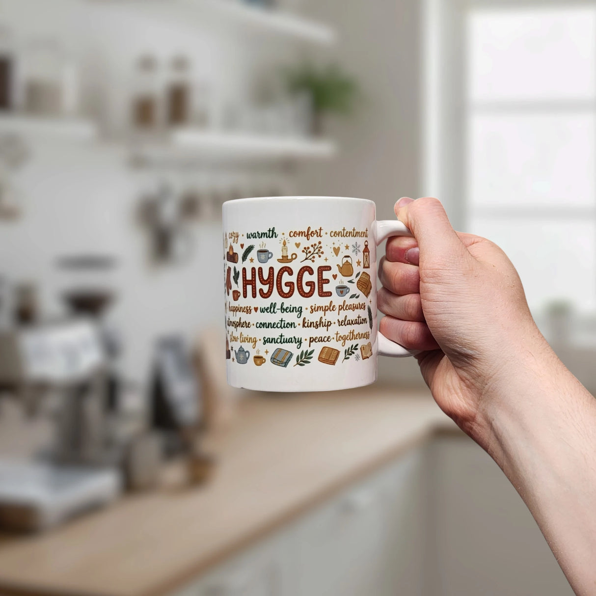 A hand holds the Swedish Gift Store's Nordic Hygge Comfort Typography Ceramic Mug (11 oz), featuring "Hygge" and cozy designs, with a bright, softly blurred kitchen in the background.