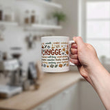 A hand holds the Swedish Gift Store's Nordic Hygge Comfort Typography Ceramic Mug (11 oz), featuring "Hygge" and cozy designs, with a bright, softly blurred kitchen in the background.