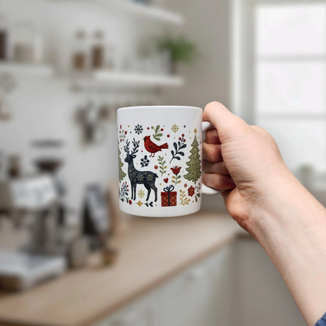 A hand holds the Nordic Wholesale Mug: Nordic Forest Reindeer & Christmas Tree Ceramic Mug (11 oz), featuring charming Scandinavian holiday motifs such as a reindeer, tree, bird, snowflakes, and gift box. A bright, blurred kitchen is visible in the background.