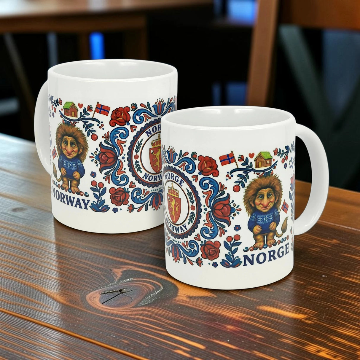 Two white 11 oz ceramic mugs by Nordic Wholesale sit on a wooden table, featuring wraparound Norwegian Rosemaling and Troll designs with lions, roses, flags, shields, and the words “Norway” and “Norge.”.