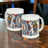 Two white 11 oz ceramic mugs by Nordic Wholesale sit on a wooden table, featuring wraparound Norwegian Rosemaling and Troll designs with lions, roses, flags, shields, and the words “Norway” and “Norge.”.