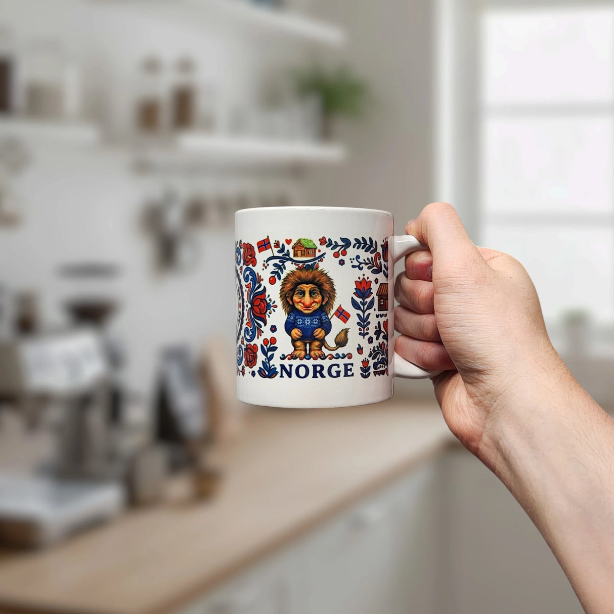 A hand holds the Nordic Wholesalers Mug: Norge Norway Rosemaling Troll Wraparound Ceramic Mug (11 oz), which features a cartoon troll, colorful Rosemaling floral patterns, and “Norge,” with a blurred kitchen in the background.
