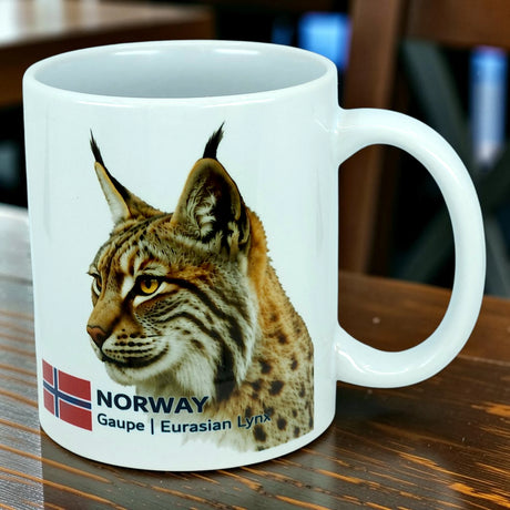 Showcased on a wooden table, the Nordic Wholesale Mug: Norway National Animal Eurasian Lynx Ceramic Mug (11 oz) displays a detailed illustration of a Eurasian lynx accompanied by the Norwegian flag and the text NORWAY Gaupe | Eurasian Lynx—a perfect celebration of Norwegian wildlife and Nordic heritage.