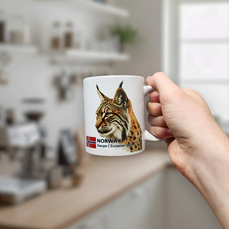 A hand holds the Nordic Wholesale Mug: Norway National Animal Eurasian Lynx Ceramic Mug (11 oz), which showcases a striking image of the majestic lynx, the word "NORWAY," and a small Norwegian flag—a tasteful tribute to Norwegian wildlife and Nordic heritage. The background reveals a softly blurred kitchen with shelves and a window.
