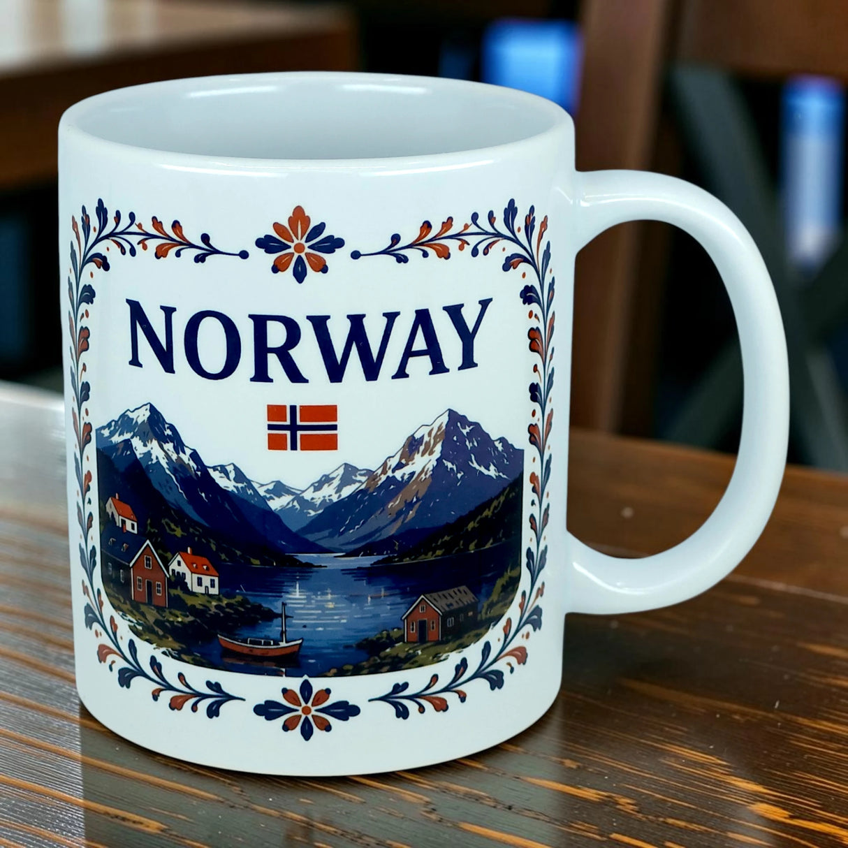 Introducing the Mug: Norway Fjord Mountain Ceramic Mug (11 oz Capacity) by Nordic Wholesale—a white mug featuring the word "Norway," the Norwegian flag, and a vibrant illustration of mountains, water, and houses. Beautifully adorned with blue and orange rosemaling-inspired floral patterns, this Norwegian heritage mug captures the timeless beauty of traditional fjord landscapes and artistry.