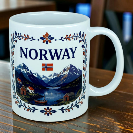 Introducing the Mug: Norway Fjord Mountain Ceramic Mug (11 oz Capacity) by Nordic Wholesale—a white mug featuring the word "Norway," the Norwegian flag, and a vibrant illustration of mountains, water, and houses. Beautifully adorned with blue and orange rosemaling-inspired floral patterns, this Norwegian heritage mug captures the timeless beauty of traditional fjord landscapes and artistry.