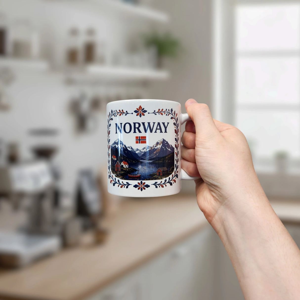 A hand holds a Nordic Wholesale Mug: Norway Fjord Mountain Ceramic Mug (11 oz Capacity), featuring white ceramic decorated with mountains, houses, and the word "NORWAY" above a small flag. A blurred kitchen with shelves and a window is visible in the background.