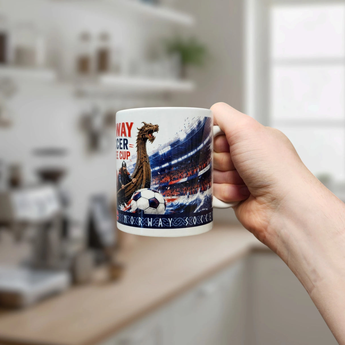A hand holds the Norway Soccer Coffee Cup Mug – Viking Longship Fan Mug by Nordic Wholesale, adorned with a Viking longship, soccer ball, and stadium scene. The mug features the NORWAY SOCCER CUP text and is set against a blurred kitchen background—a perfect Norwegian gift for soccer enthusiasts.