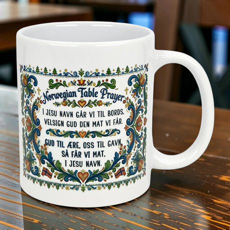 The Nordic Wholesale Mug: Norwegian Table Prayer Ceramic Rosemaling Mug (11 oz) features vibrant floral borders and the prayer in Norwegian and English, making it a perfect heritage gift. The white ceramic mug with ornate blue-green patterns sits on wood.