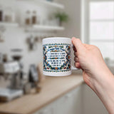 A person holds a white Nordic Wholesale Rosemaling mug with an intricate, colorful border and “Norwegian Table Prayer” text. In the background is a blurred kitchen with shelves, jars, and a coffee machine.