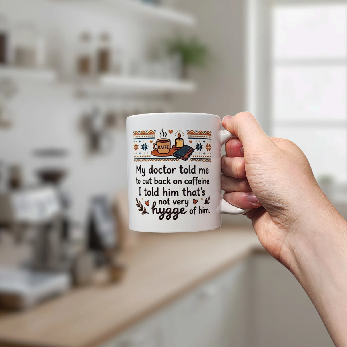 A hand holds the Swedish Gift Store’s "Not Very Hygge" Coffee Mug (11 oz) in a kitchen. The white mug features coffee and cozy-themed illustrations above the text: My doctor told me to cut back on caffeine. I told him that’s not very hygge of him.