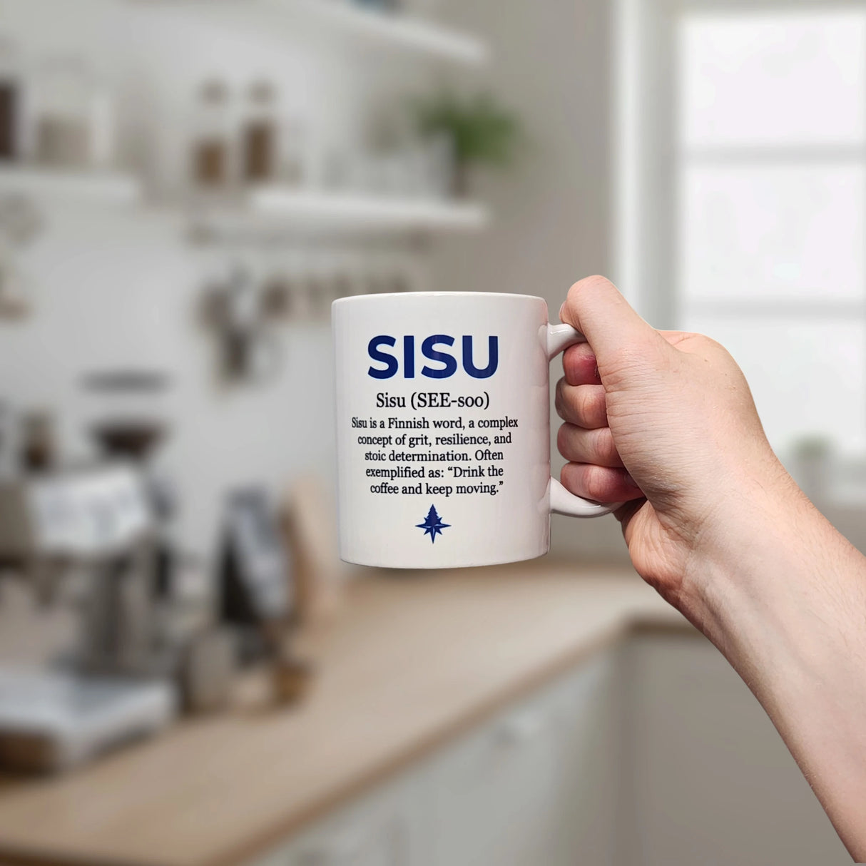 A hand holds the Nordic Wholesalers Mug: Sisu Finnish Word Definition Ceramic Mug (11 oz) in a kitchen. The white mug features “SISU” with its pronunciation and definition—highlighting Finnish grit, resilience, and determination. Blurred shelves are behind.