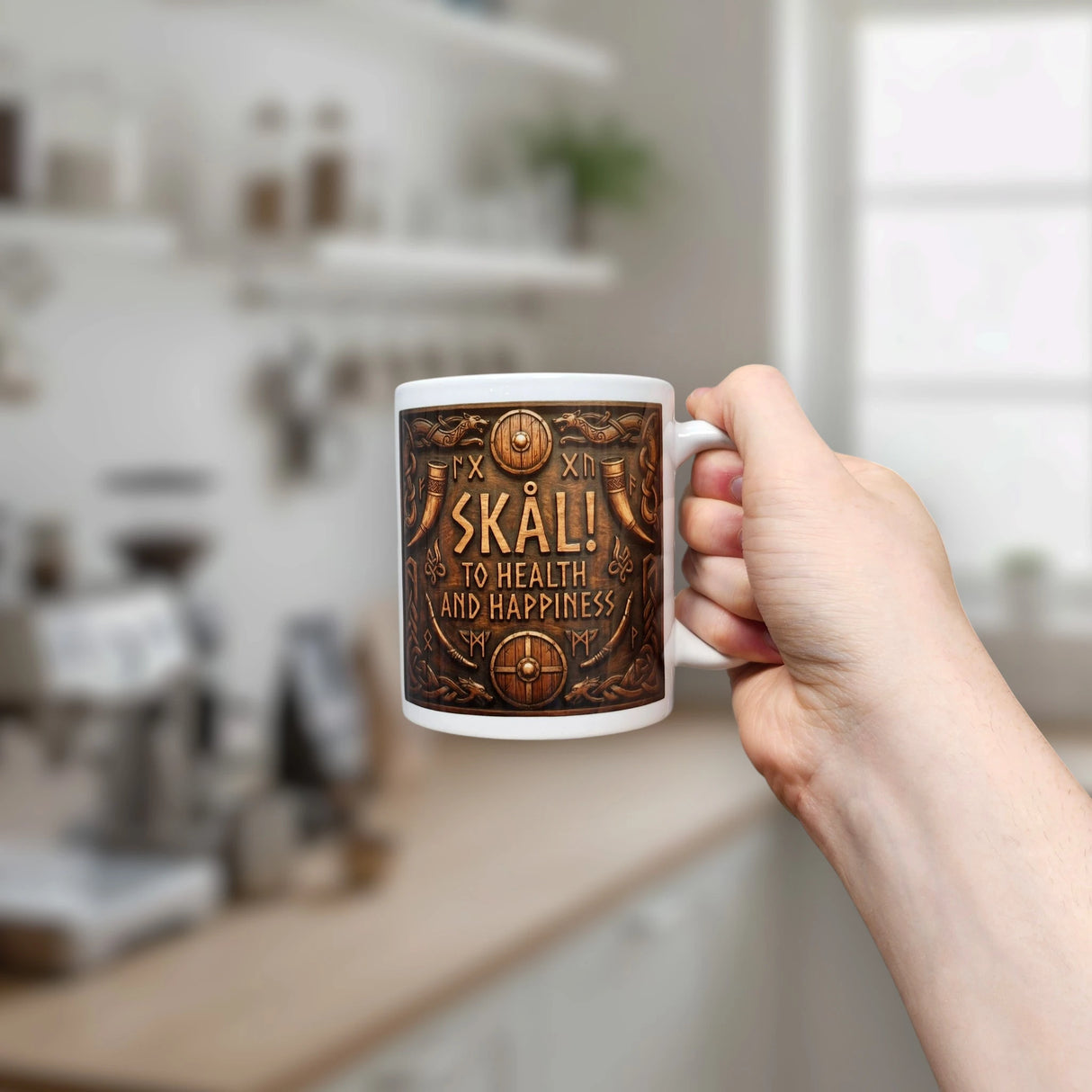 A hand holds the Nordic Wholesale Mug: Skal To Health And Happiness Norse Ceramic Mug (11 oz), which features the phrase “SKÅL! TO HEALTH AND HAPPINESS” in a kitchen with blurred shelves, plants, and coffee equipment in the background.