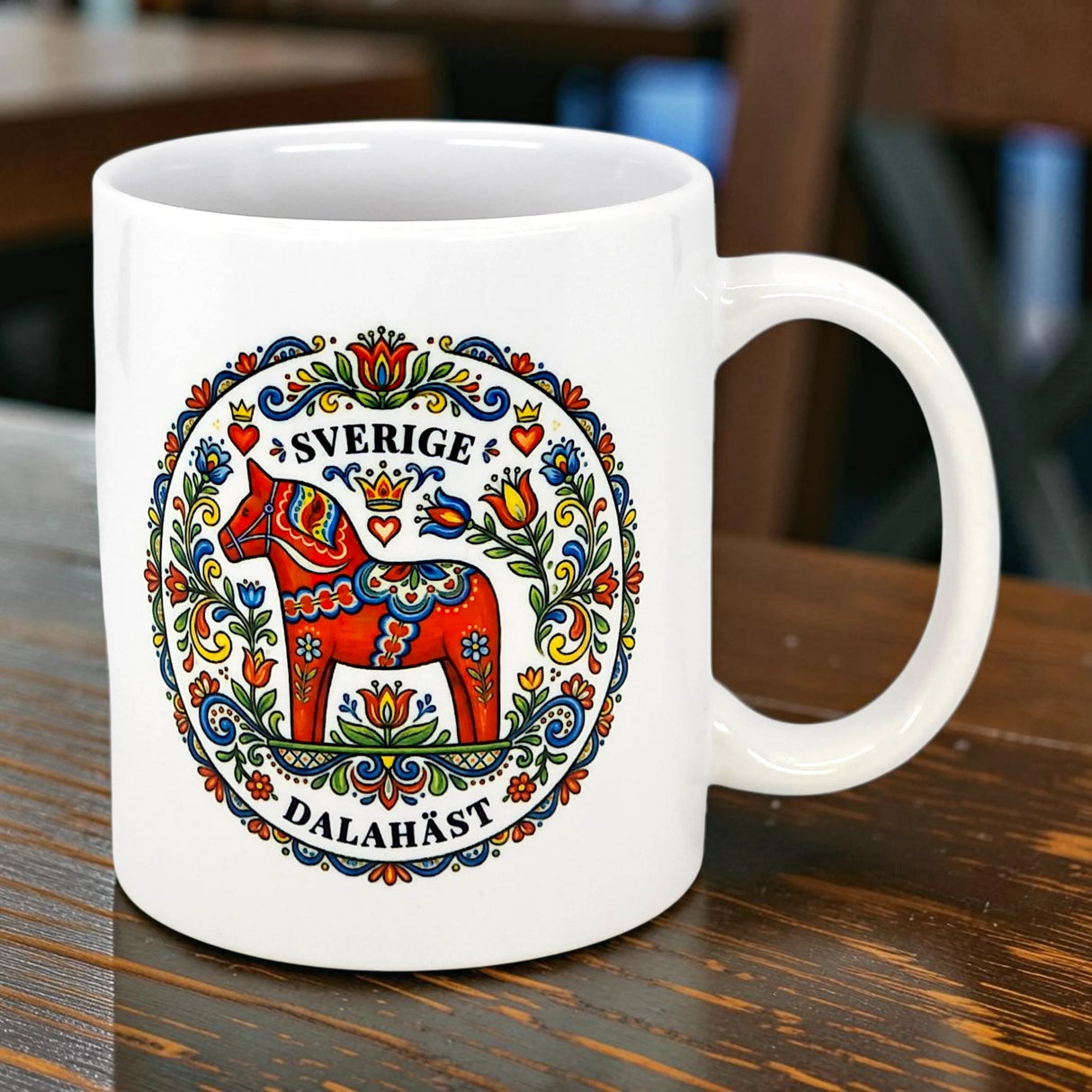 The Swedish Gift Store "Sverige Dala Häst" 11oz ceramic mug features a red Dala horse with a Swedish folk art wreath and the words Sverige and Dalahäst on the front, shown on a wooden table.