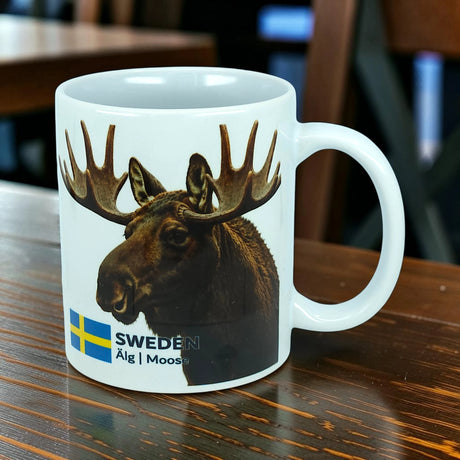 The Nordic Wholesale Mug: Sweden Älg Moose Ceramic Mug (11 oz) features a white ceramic design adorned with a photo of a moose, the Swedish flag, and the text "SWEDEN Älg | Moose." Placed on a wooden table, this charming mug is the perfect addition to any Scandinavian kitchen and beautifully celebrates Nordic nature.