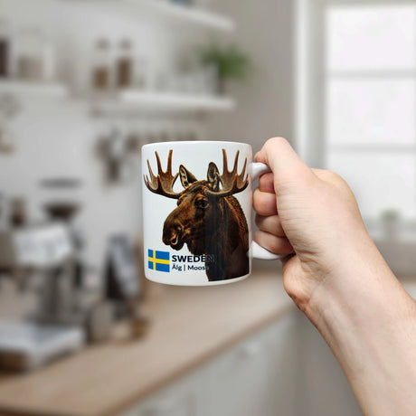 In a bright, Scandinavian-inspired kitchen that reflects the essence of Nordic nature, a hand holds an 11 oz white ceramic "Mug: Sweden Älg Moose" by Nordic Wholesale, featuring an image of a moose, the Swedish flag, and the words “SWEDEN Älg | Moose.”.