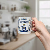 A hand holds the Nordic Wholesale Mug: Sweden Dala Horse Ceramic Mug (11 oz Capacity), featuring blue and yellow patterns, a Swedish flag, the word "Sweden," and a horse illustration. The background displays a blurred kitchen.