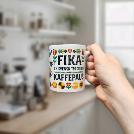 A hand holds the Nordic Wholesale "Fika — En Svensk Tradition Kaffepaus" Swedish Fika Traditions Wraparound Ceramic Mug (11 oz), decorated with flowers, hearts, and cookies, framed by a bright, blurred kitchen background.