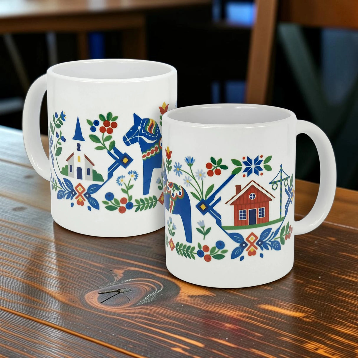 Two "Swedish Folk Art — Blue Dala Horse, Church & Red House" 11oz ceramic mugs by Nordic Wholesale, featuring Scandinavian folk art designs in blue, red, green, and orange with a Dala Horse, flowers, leaves, a church, and a red house, are placed on a wooden table.