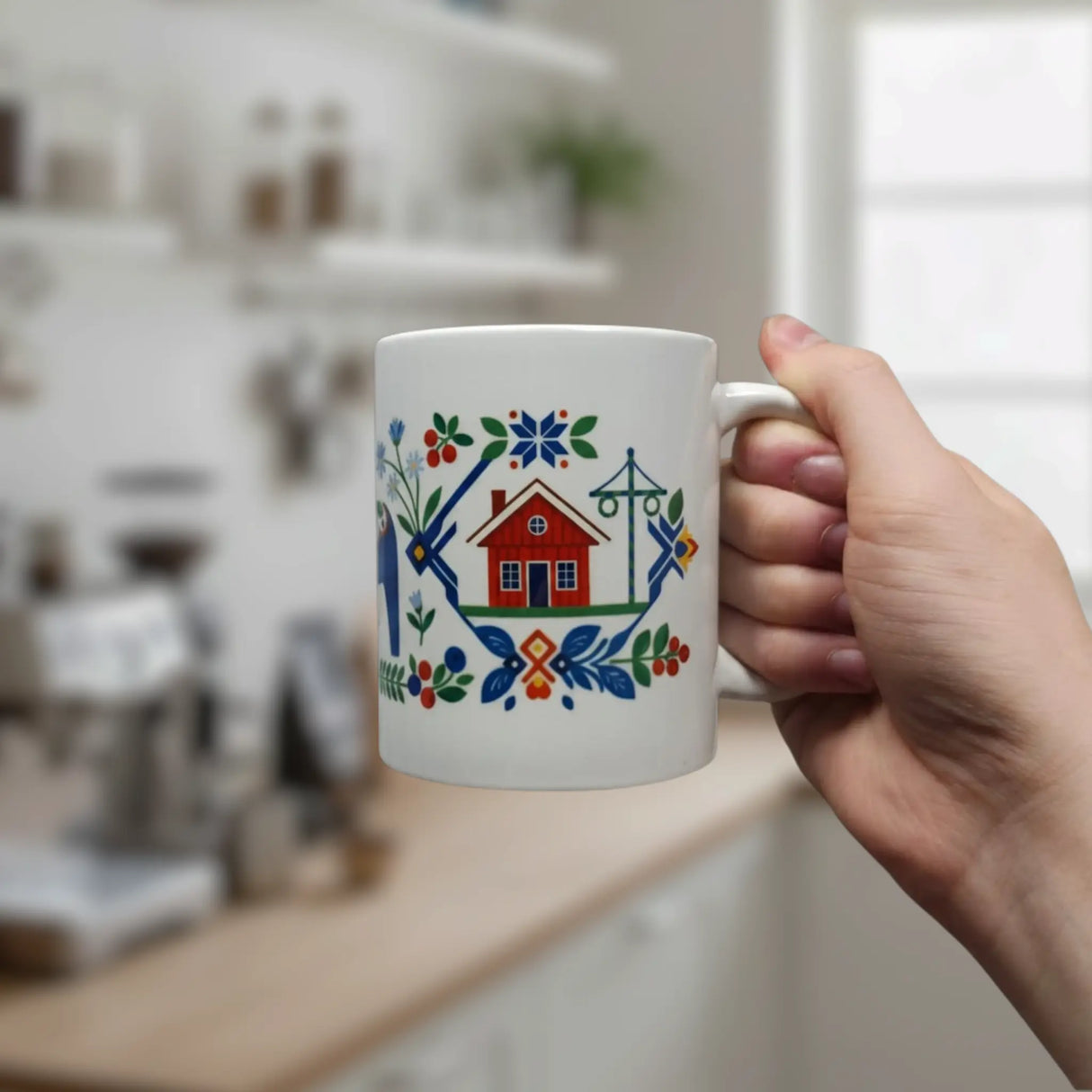 A hand holds a Nordic Wholesale 11oz ceramic mug, featuring the Mug: Swedish Folk Art design with a blue Dala horse, church, red house, trees, and vibrant floral patterns. The background shows a softly blurred kitchen.