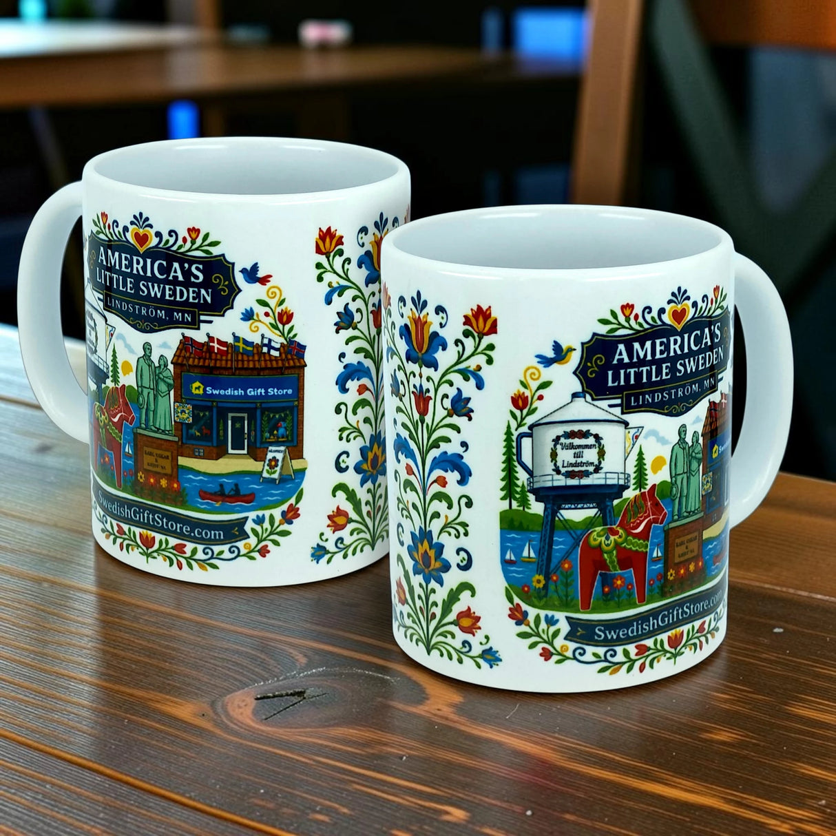 Two white Nordic Wholesale ceramic mugs (11 oz), featuring colorful floral patterns and shop illustrations, display the text "America’s Little Sweden, Lindstrom, MN" and "SwedishGiftStore.com." The mugs from the Swedish Gift Store collection rest on a wooden table.