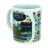 Introducing the Mug: Swedish Gift Store America's Little Sweden Ceramic Mug (11 oz) by Nordic Wholesale. This white ceramic mug showcases vibrant Swedish folk art, a storefront with the “Swedish Gift Store” sign, and a statue, along with text that reads “America’s Little Sweden, Lindström, MN.” The handle and right side are adorned with charming floral patterns and a cheerful Dala horse design for an authentic Nordic touch.
