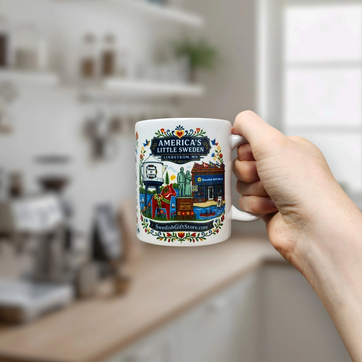 A hand holds the Nordic Wholesale Mug: Swedish Gift Store America's Little Sweden Ceramic Mug (11 oz), featuring colorful artwork and text reading “America’s Little Sweden, Lindsborg, KS,” in a bright kitchen setting.