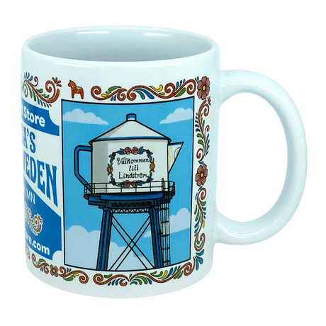 The Nordic Wholesale "Mug: Swedish Gift Store America’s Little Sweden Wraparound Ceramic Mug (11 oz)" features a charming illustration of a teapot-shaped water tower with “Välkommen till Lindström” displayed on it, surrounded by floral borders and set against a blue sky with clouds—a delightful tribute to Swedish-American heritage.