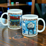 Two white ceramic mugs with vibrant Scandinavian motifs rest on a wooden table. One, the Nordic Wholesale 11 oz "Swedish Gift Store America's Little Sweden" wraparound ceramic mug, features a charming illustration of a gift store, while the other displays a water tower labeled "Little Sweden.