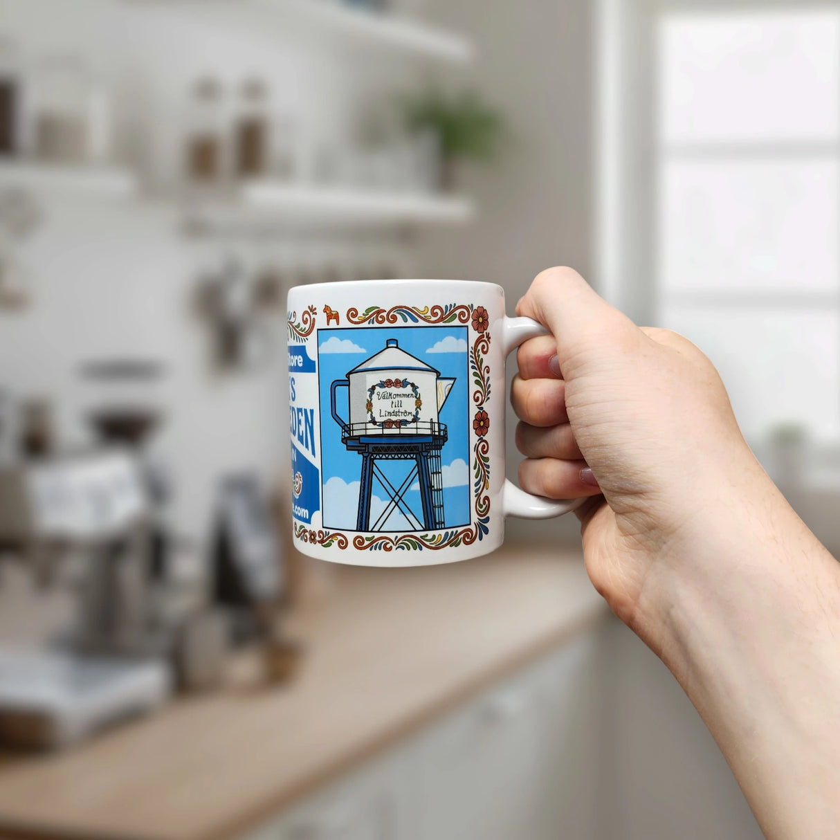 A hand holds the Nordic Wholesale "America's Little Sweden" Wraparound Ceramic Mug (11 oz), featuring decorative borders and a coffee pot-topped water tower illustration, with text celebrating Swedish-American heritage, set against a bright, blurred kitchen background.