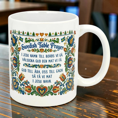 The Nordic Wholesale Mug: Swedish Table Prayer Ceramic Kurbits Mug (11 oz) features a white base with a colorful Scandinavian floral Kurbits design and the Swedish Table Prayer—perfect as a heritage gift, shown on a wooden table.