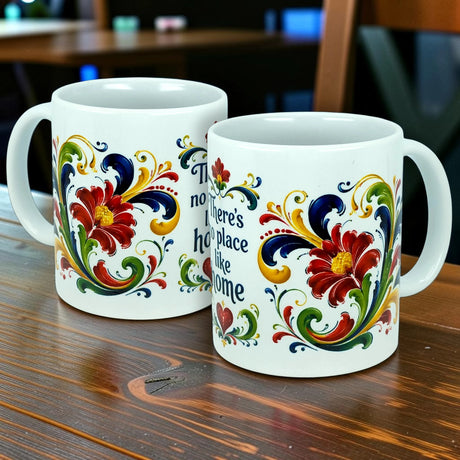 Two 11 oz white ceramic mugs from Nordic Wholesale, featuring colorful Scandinavian rosemaling patterns and the phrase "There's No Place Like Home," sit on a wooden table.