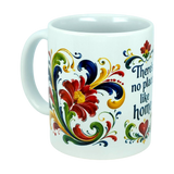 The Mug: There's No Place Like Home Rosemaling Ceramic Mug (11 oz) by Nordic Wholesale is a white ceramic coffee mug featuring red, yellow, blue, and green floral designs inspired by Scandinavian folk art. Ornate swirls reminiscent of traditional Norwegian Rosemaling surround the flowers, and the partial phrase "There's no place like home" appears in blue text on the mug.