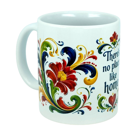 The Mug: There's No Place Like Home Rosemaling Ceramic Mug (11 oz) by Nordic Wholesale is a white ceramic coffee mug featuring red, yellow, blue, and green floral designs inspired by Scandinavian folk art. Ornate swirls reminiscent of traditional Norwegian Rosemaling surround the flowers, and the partial phrase "There's no place like home" appears in blue text on the mug.