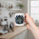 A hand holds the Nordic Wholesale “This Is My Raiding Mug” Viking Ceramic Mug (11 oz), featuring dark fantasy artwork and text, in a kitchen with blurred shelves and a window in the background.
