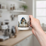 A hand holds the Nordic Wholesale Mug: Troll Fuel Ceramic Coffee Mug (11 oz), showing a bearded troll and the text "Troll Fuel. This coffee is mine. Trespassers will be caffeinated." A bright, blurred kitchen appears in the background.