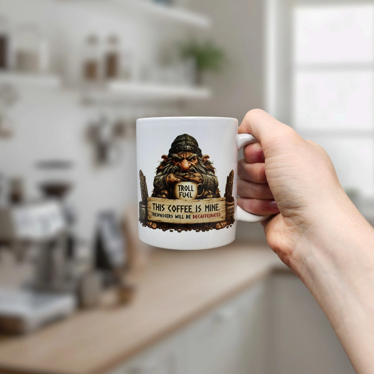 A hand holds the Mug: Troll Fuel Ceramic Coffee Mug (11 oz Capacity) by Nordic Wholesale—a white ceramic mug decorated with a cartoon troll and the playful warning, “Troll Fuel. This coffee is mine. Trespassers will be decaffeinated,” perfectly capturing the whimsical spirit of Scandinavian folklore in a bright kitchen setting.