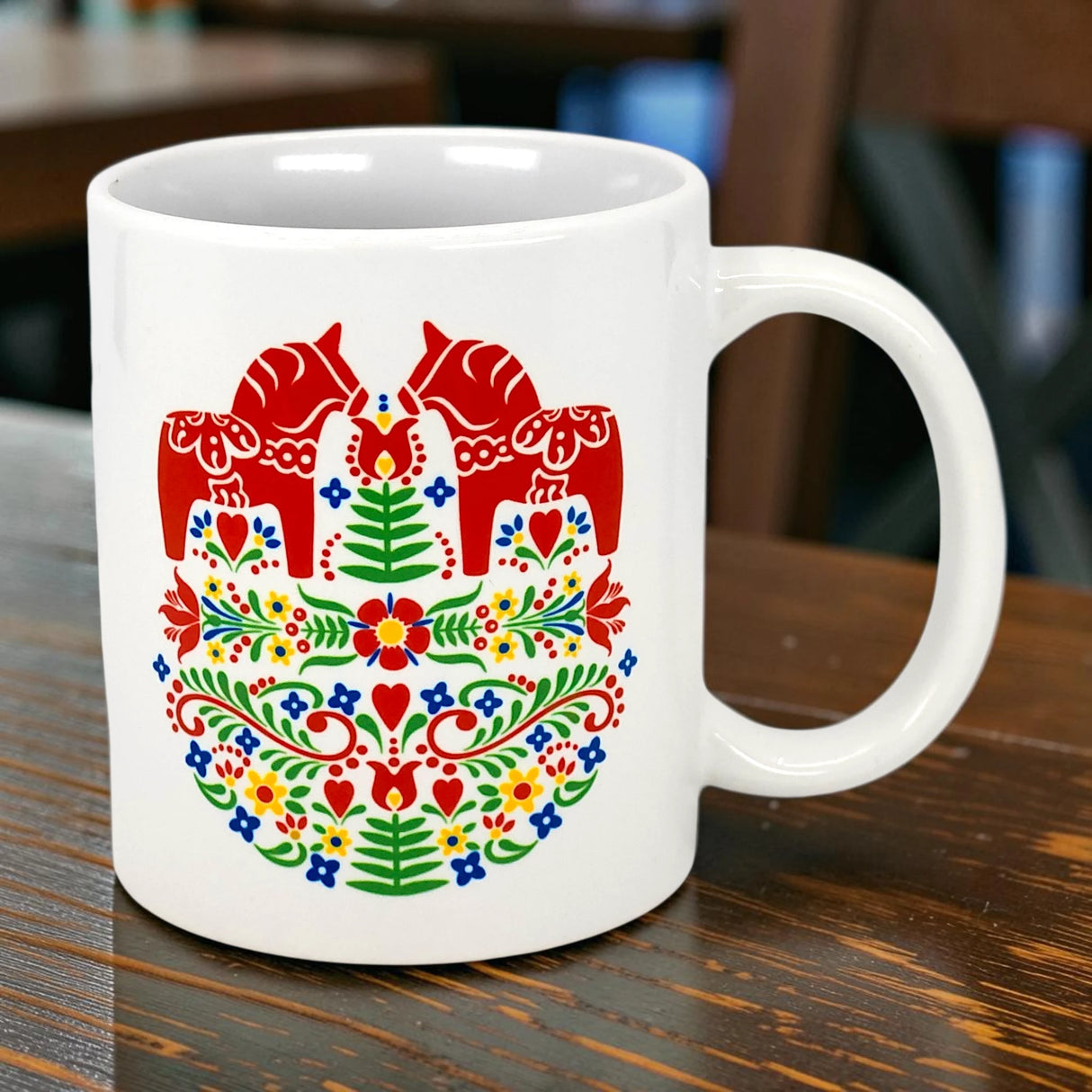 The 11oz ceramic "Two Red Dala Horses in a Field of Swedish Folk Flowers" mug from Swedish Gift Store features vibrant Scandinavian folk art with red Dala horses, green leaves, red hearts, and colorful flowers.
