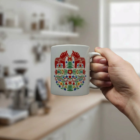 A hand holds the Nordic Wholesale 11oz ceramic mug, featuring two red Dala horses amid Swedish folk flowers in a colorful pattern; a blurred kitchen appears in the background.