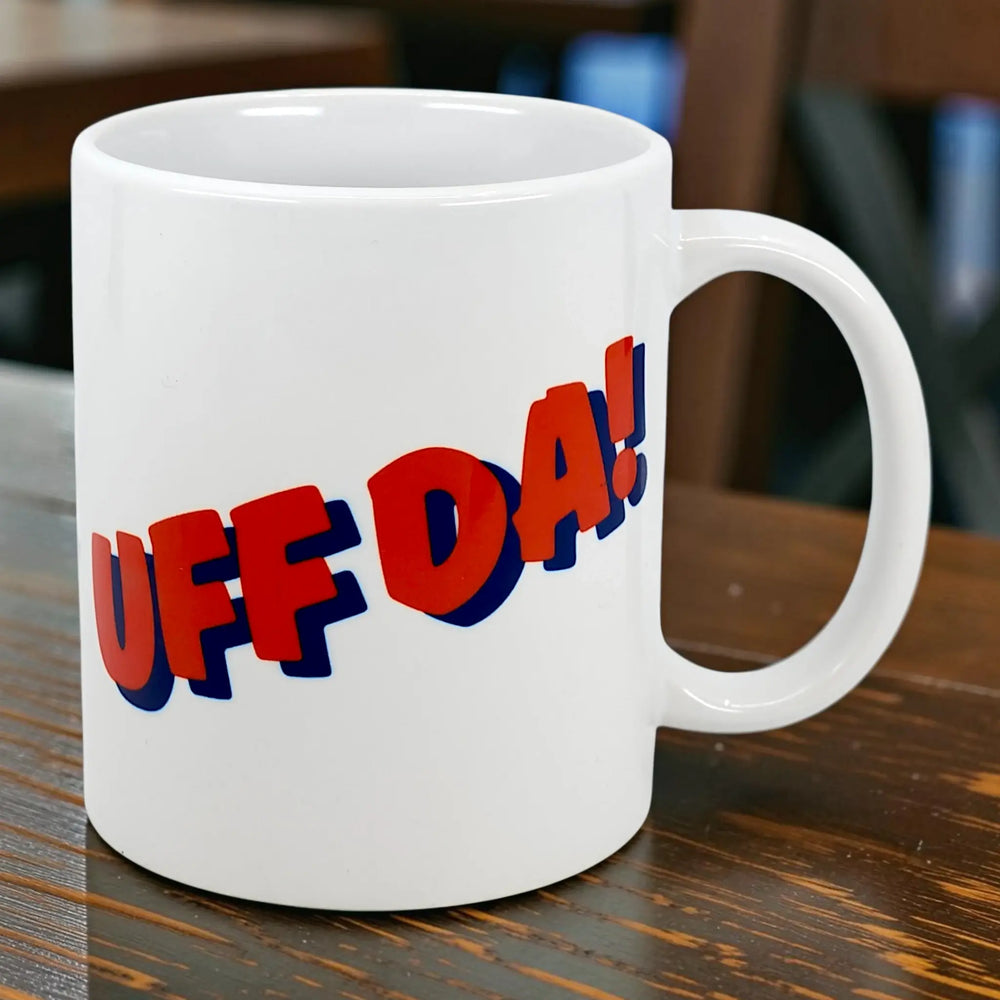 The Mug: Classic "Uff Da!" Ceramic Mug (11 oz) by Nordic Wholesale sits on a wooden table, showcasing bold blue and red 3D text. The standout Scandinavian mug contrasts with a blurred background of chairs and tables.