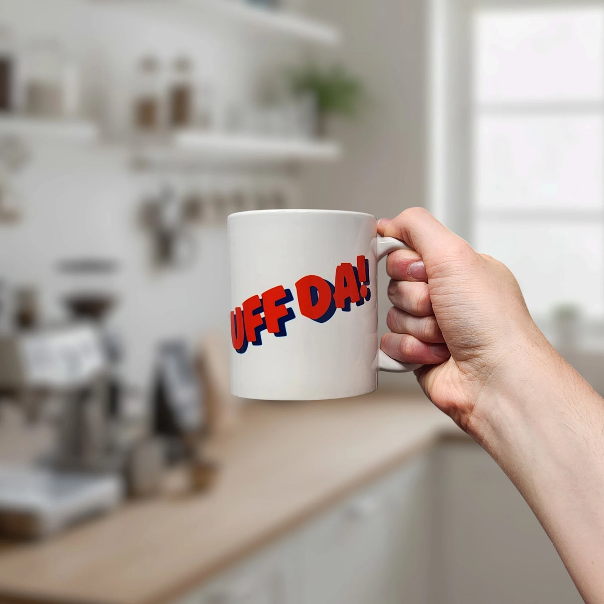 A hand holds the Swedish Gift Store’s Classic "Uff Da!" Ceramic Mug (11 oz), a white cup with bold red and blue “UFF DA!” lettering; the blurred kitchen background shows shelves, jars, and a coffee machine.
