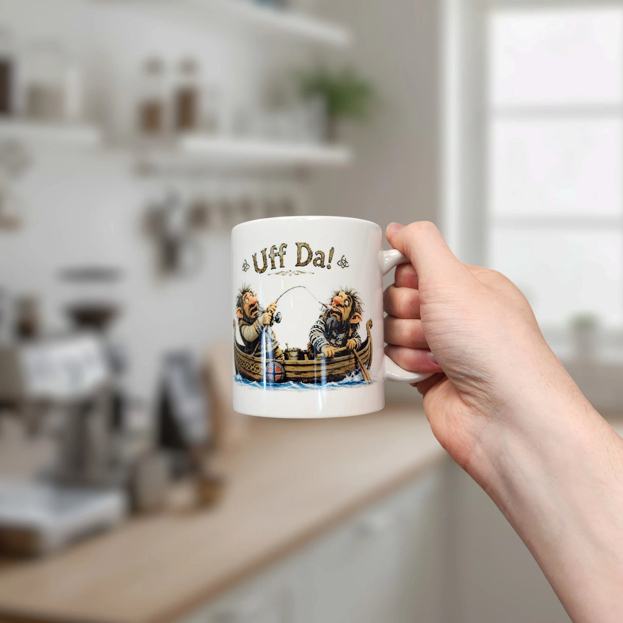 A hand holds the Nordic Wholesale Mug: Uff Da Fishing Trolls Ceramic Mug (11 oz), featuring two people in a boat under the phrase “Uff Da!” amid a cozy, blurred kitchen and window backdrop.