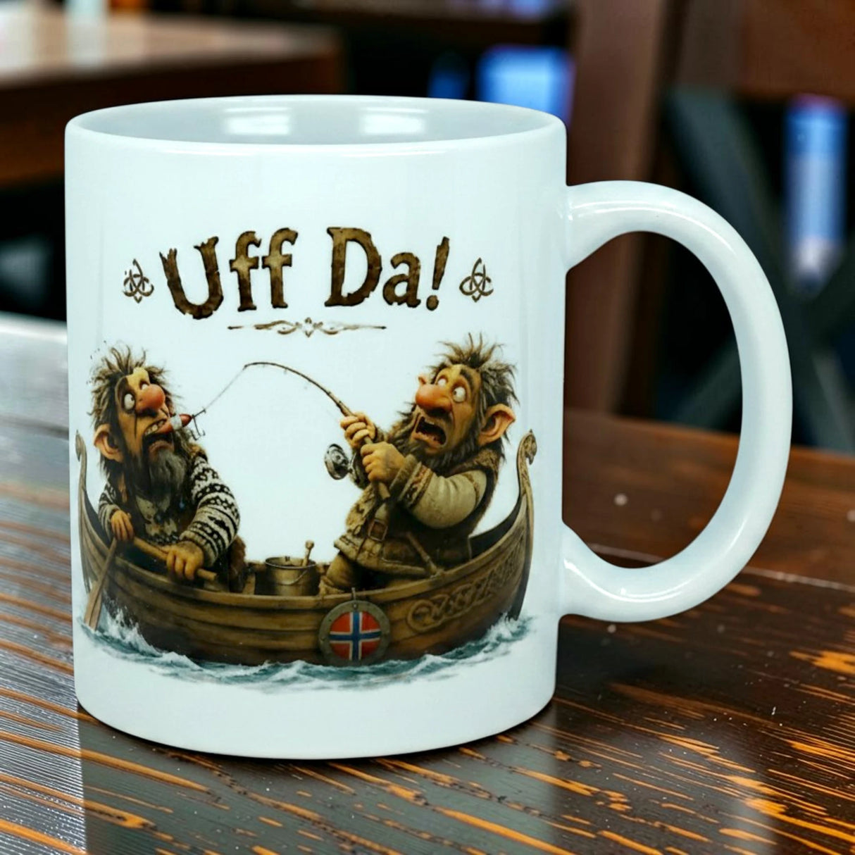 The Nordic Wholesale Mug: Uff Da Fishing Trolls Ceramic Mug (11 oz Capacity) showcases two cartoon trolls fishing from a wooden boat, with "Uff Da!" displayed above them and a small Norwegian flag on the boat. This Scandinavian humor mug is pictured against a softly blurred indoor background.