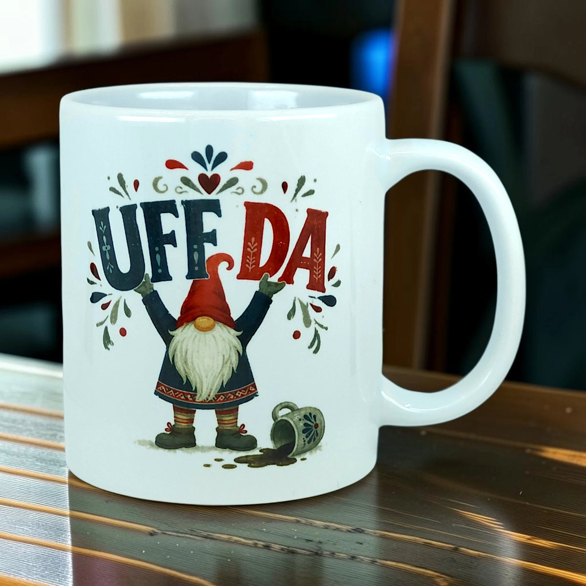 White ceramic mug featuring a tomte gnome with a red hat and white beard standing next to a spilled mug, with the text UFF DA in blue and red.