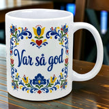 White mug with “Var Så God” in blue script and colorful floral patterns inspired by Swedish folk art on the side, placed on a wooden table.