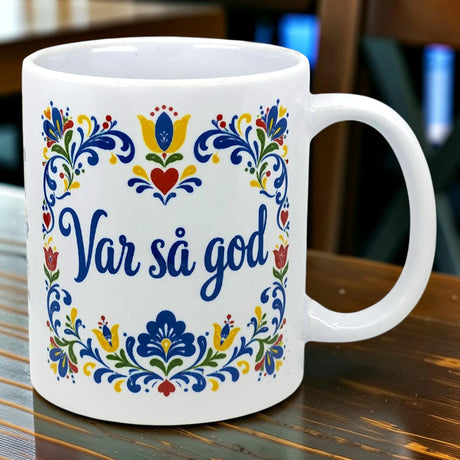 White mug with “Var Så God” in blue script and colorful floral patterns inspired by Swedish folk art on the side, placed on a wooden table.