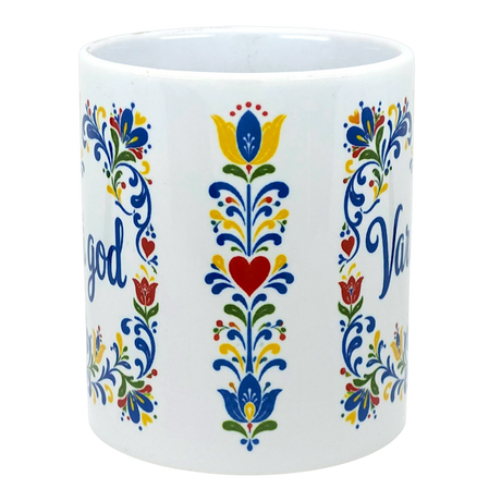 White ceramic Scandinavian mug featuring Swedish folk art patterns of flowers and hearts in red, yellow, blue, and green. Partial text “Var Så God” is visible.