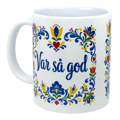 White mug featuring Swedish folk art with multicolored floral motifs surrounding blue text “Var så god” centered on the front.